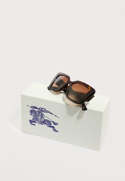 Γυαλιά ηλίου υπερμεγέθη τετράγωνα με φιλοτεχνημένο μοτίβο χελώνας και καφέ φακούς που ακουμπούν πάνω σε ένα λευκό κουτί με έμβλημα μπλε ιππότη επάνω σε άλογο.