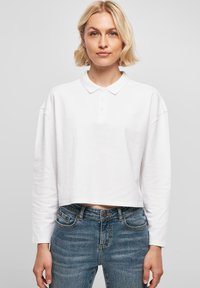 Weißes, kurz geschnittenes Langarm-Polohemd mit Kragen und einer Drei-Knopf-Leiste, kombiniert mit blauen Jeans aus Denim, die über Fronttaschen verfügen.