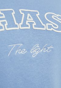 Lichtblauwe trui met opvallende witte letters met een gestructureerde omlijning, en een subtiele ingenaaide tekst eronder in cursief.