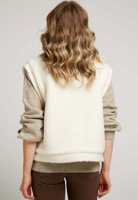 Veste en polaire crème par-dessus un top à manches longues beige, avec des manches roulées et un ourlet marron. Pantalons marron visibles en bas. Surface texturée.