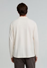 Cămașă polo cu mânecă lungă, de culoare albă murdară, cu o textură moale, guler și tiv drept. Purtați-o cu pantaloni din catifea cotată maro închis.