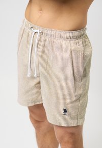 Beige és fehér csíkozott strandnadrág, rugalmas derékkal, állítható húzózsinórral és oldalsó zsebekkel. Egy kis kék logóval rendelkezik.
