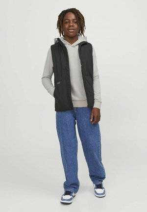 Zwarte mouwloze vest over een lichtgrijze hoodie, gecombineerd met losse blauwe jeans en witte sneakers met blauwe accenten. Twee voorzakken zichtbaar.