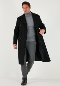 Cappotto lungo in lana nera con design a doppio petto, grandi rever e due bottoni, indossato sopra un dolcevita grigio e pantaloni grigi.