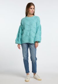 Aquamarinfarbener oversized Pullover mit texturiertem wellenförmigem Muster, geripptem Hals und Bündchen, kombiniert mit hellblauen Jeans und weißen Sneakern.