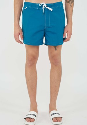 Mannelijke benen met blauwe zwembroek met witte trekkoord en witte slippers op een effen witte achtergrond.