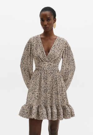 Vrouw draagt een beige jurk met zwarte vlekken, diepe V-hals, lange pofmouwen, getailleerde taille en gerimpelde zoom, tegen een effen achtergrond.