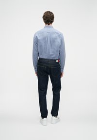 Άνδρας που στέκεται με πλάτη, φορώντας μπλε ριγέ πουκάμισο, σκούρες τζιν παντελόνες με λογότυπο και λευκά αθλητικά παπούτσια, με φόντο λευκό.