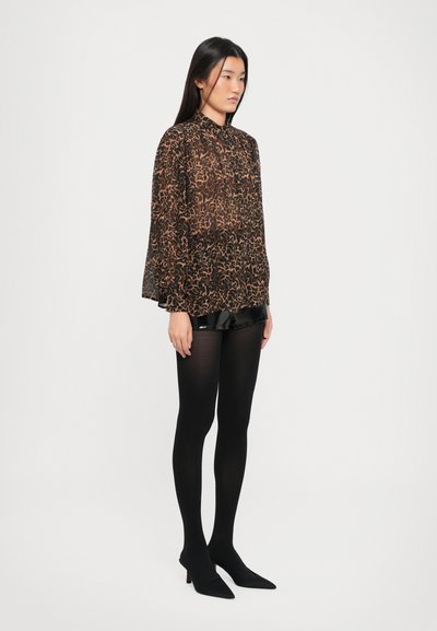 Sort gennemsigtig blouse med abstrakt brunt mønster, lange ærmer og høj krave, parret med skinnende sorte shorts og sorte strømpebukser, som fuldender outfittet med spidse hæle.