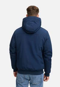 Hombre con una chaqueta con capucha azul marino y vaqueros azules, mostrado desde atrás contra un fondo blanco liso.