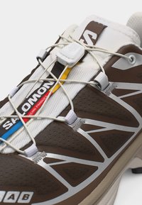 Κοντινό πλάνο καφέ και λευκού παπουτσιού Salomon με ελαστικό σύστημα γρήγορου κορδονιού και πολύχρωμη επωνυμία στη γλώσσα.