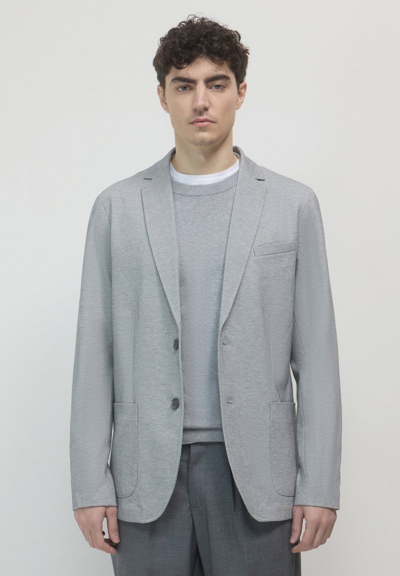 Blazer grigio chiaro in tessuto texturizzato, con due tasche frontali e rever a punta, indossato sopra un maglione grigio chiaro coordinato.
