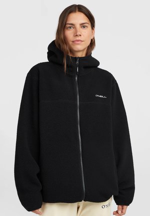 Vrouw draagt een zwarte fleece zip-hoodie met O'Neill-logo op de borst en beige broek met logo op de dij tegen een effen achtergrond.
