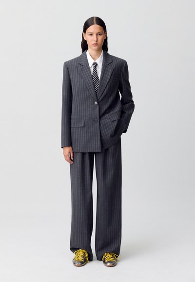 Traje gris a rayas con blazer oversized y pantalones de pierna ancha. Combinado con una camisa blanca y una corbata a rayas blanca y negra. Zapatillas amarillas con cordones.