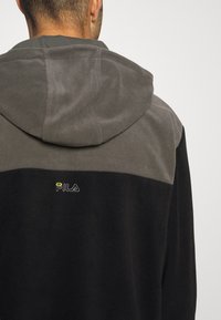 Sort og grå hættetrøje med en blød tekstur. Har et farveblokdesign med kontrast og et broderet "FILA" logo i gul.