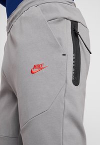 Joggers grises de Nike con un bolsillo con cremallera, que presentan un logo rojo bordado y costuras texturizadas. Un diseño elegante y ceñido mejora el ajuste.