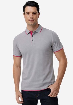 Ein Mann trägt ein eng anliegendes graues Poloshirt mit pinkem Besatz am Kragen, an der Knopfleiste und an den Ärmelenden, kombiniert mit dunkelblauen Jeans.