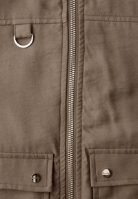 Close-up af brun stofjakke med central sølvlynlås, D-ring på venstre side og to lommer med klapper og knapper nederst.