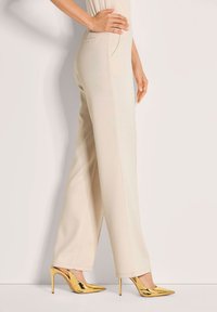 Vrouw in crèmekleurige hooggetailleerde broek, gecombineerd met glanzende gouden puntige stilettohakken, staand tegen een lichte achtergrond.