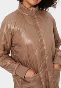 Veste matelassée en duvet de couleur marron clair, avec un col haut, une fermeture éclair complète, des poignets côtelés et des poches latérales. Texture lisse et brillante avec un motif en losanges.