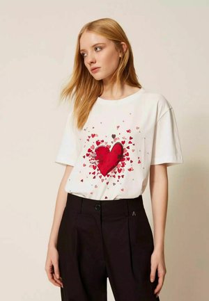 Donna con capelli biondi lunghi che indossa una maglietta bianca con un grande cuore rosso e piccoli cuori sparsi, abbinata a pantaloni neri.