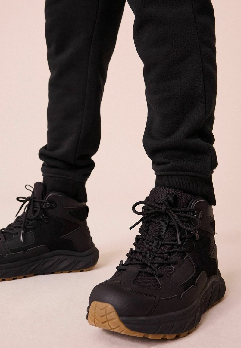Czarne sneakersy typu high-top z teksturowanej tkaniny i gumowymi akcentami. Posiadają mocne sznurówki, wyściełaną podeszwę oraz kontrastującą gumową podeszwę.