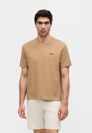 Férfi, aki texturált, bézs rövid ujjú pólót visel "BOSS" logóval és világos rövidnadrággal, áll egy egyszínű fehér háttér előtt.