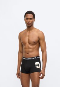 Juodos bokso šortai su balta Karl Lagerfeld logotipu ant juosmens juostos ir animacinio personažo grafika priekyje. Medvilnės medžiaga, prigludęs siluetas.