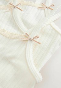 Světle béžová tílko vyrobené z ribbed látky, s jemným krajkovým lemem a malými mašličkami na výstřihu a ramínkách.