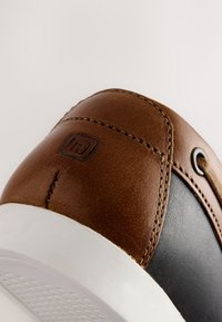 Gros plan du talon d'une basket en cuir marron avec des détails cousus et un panneau latéral noir au-dessus de la semelle en caoutchouc blanche.