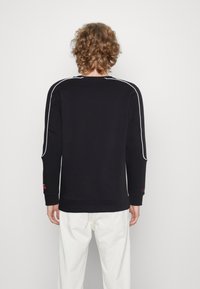 Sudadera negra con piping blanco a lo largo de los hombros y las mangas. Puños y dobladillo de canalé, con detalle del logotipo en la manga. Vista trasera lisa.