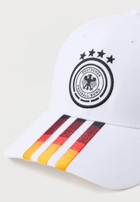 Λευκό καπέλο μπέιζμπολ με το λογότυπο της Γερμανικής Ομοσπονδίας Ποδοσφαίρου και τέσσερα αστέρια από πάνω, με μαύρες, κόκκινες και κίτρινες ρίγες γύρω από την άκρη.