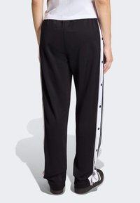 Crne sportske hlače s bijelim bočnim prugama i detaljima sa kopčama, izrađene od glatkog materijala, s opuštenim krojem i elastičnim strukom.
