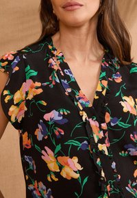 Un haut noir à motifs floraux présente un décolleté en V, des manches courtes froncées et des motifs floraux vifs en jaune, rose et bleu sur un fond sombre.