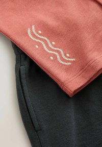 Korbrotmischung Oberteil in Koralle mit weißen Wellen- und Punktstickereien, geschichtet über dunkelgrauen Hosen mit weicher Textur und tailliertem Design.
