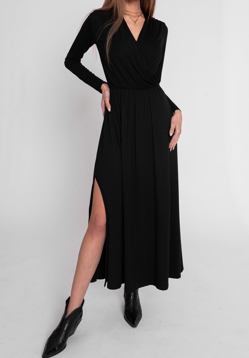 Femme en longue robe noire à manches longues et fente haute, posant avec une main sur la hanche, portant des bottines noires à talons pointus et un collier en croix.