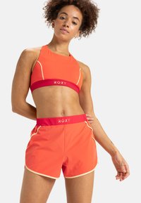 Felkleurige oranje sportbeha en bijpassende korte broek. De korte broek heeft een rode tailleband met het "ROXY"-logo en contrasterende crèmekleurige piping. Gladde stofstructuur.