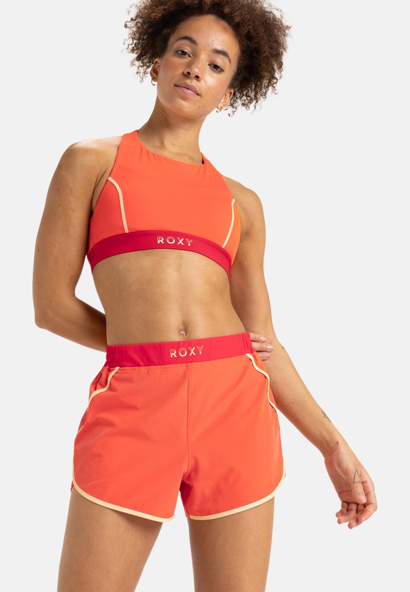 Felkleurige oranje sportbeha en bijpassende korte broek. De korte broek heeft een rode tailleband met het "ROXY"-logo en contrasterende crèmekleurige piping. Gladde stofstructuur.