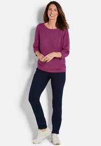 Lila Strickpullover mit Rundhalsausschnitt und Dreiviertelärmeln, kombiniert mit marineblauen Jeans und weißen Sneakers. Strukturmuster über das gesamte Kleidungsstück.
