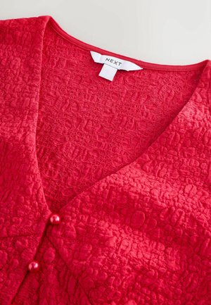 Cardigan roșu texturat cu decolteu în V, două nasturi lucioși și modele florale detaliate. Eticheta spune "NEXT." Finisaj din țesătură moale.
