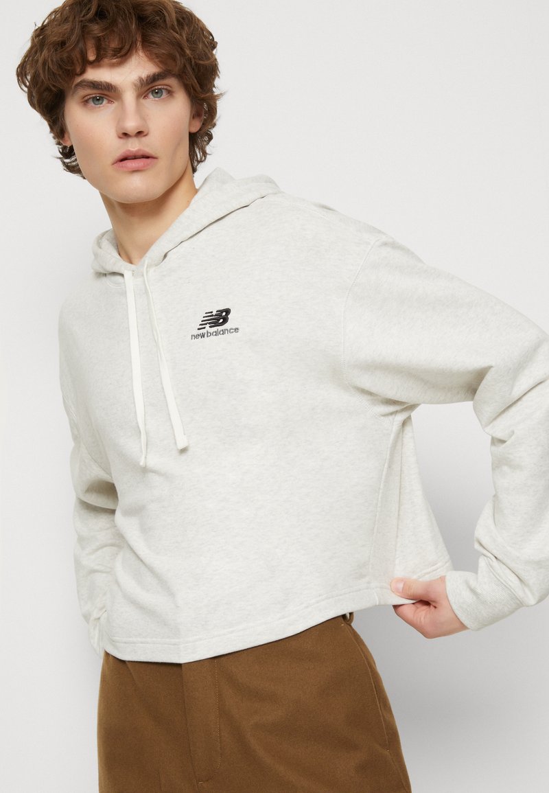 Haut à capuche cropped gris clair avec cordon de serrage et logo noir ; poignets côtelés ; texture douce ; associé à un pantalon marron.