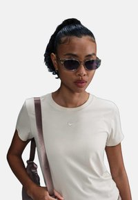 T-shirt beige a maniche corte con il logo Nike bianco, abbinato a occhiali da sole trasparenti e una borsa a tracolla marrone scuro con una tracolla in satin.