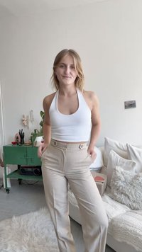 Bela halter crop majica v kombinaciji z bež hlačami visoke pasu, ki imajo dva sprednja gumba. Gladka tekstura, prilegajoč kroj.