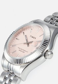 Montre en acier inoxydable argenté avec une lunette striée, un cadran rose et des index horaires argentés. Présente un bracelet lisse et une couronne texturée.