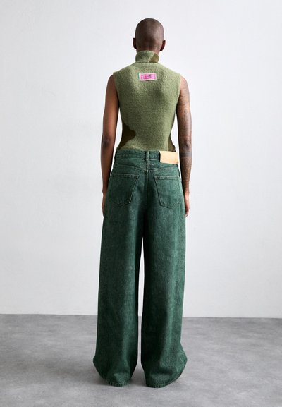 Πράσινη αμάνικη μπλούζα από υφασμάτινο υλικό, συνδυασμένη με φαρδιά πράσινα τζιν. Σημαντική ροζ ετικέτα στο πίσω μέρος της μπλούζας.