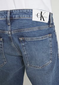 Pantalones vaqueros de mezclilla en un lavado azul medio con corte recto, con bolsillos traseros y una etiqueta blanca de Calvin Klein en la cintura.