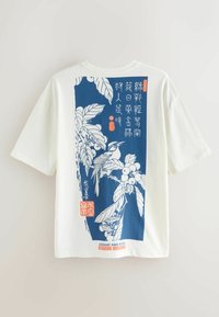 Fehér póló kék téglalap alakú nyomattal, amely egy madarat ábrázol loquat ágakon, levelekkel, gyümölcsökkel és japán stílusú szöveggel.