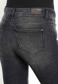 Jeans de mezclilla gris oscuro con un diseño ajustado. Las características incluyen dos bolsillos traseros y una etiqueta de cuero marrón con el nombre de la marca.