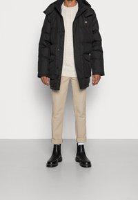 Svart pufferjacka med huva, tjock vaddering och två fickor fram, bärs över en krämfärgad stickad tröja, beige byxor och svarta ankelboots.