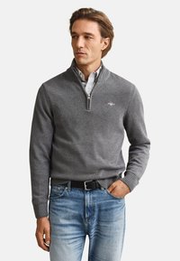 Mann trägt einen grauen Pullover mit Viertelreißverschluss über einem gestreiften Hemd, blaue Jeans und schwarzen Gürtel, eine Hand in der Tasche.